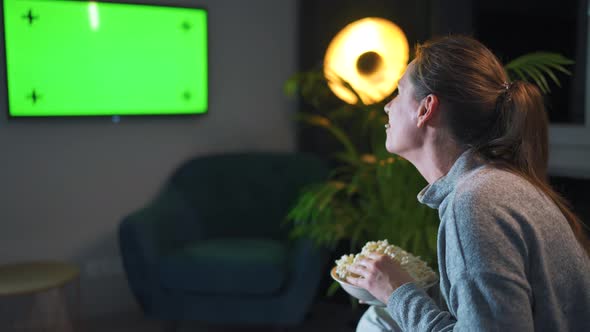 Rear View of a Woman Sitting on a Sofa in the Living Room in the Evening and Watching a Green TV