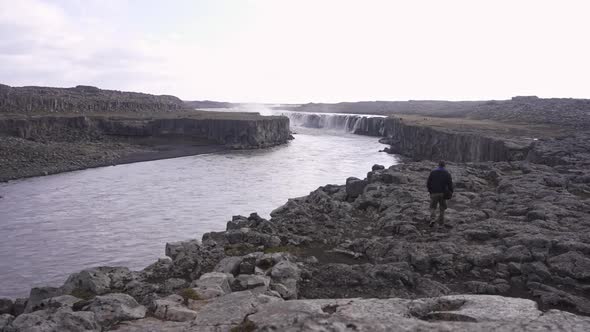 Traveler walking along rocky shore of river towards waterfall