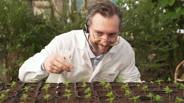 Mad Scientist in White Coat and Glasses Sprays Plant Seedlings with Water From Spray Gun