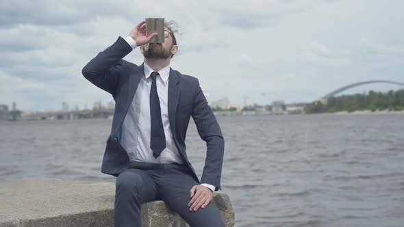 Depressed Man in Eyeglasses Drinking From Flask on River Bank with Blurred City at the Background