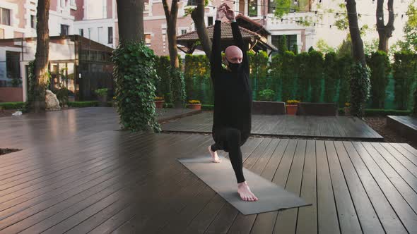 Flexible Bald Man in Black Protective Mask and Sportswear is Practicing Yoga on Mat in Courtyard