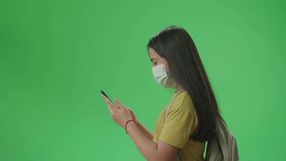 Side View Of Asian Girl Student Wearing A Mask And Use Mobile Phone While Walking On Green Screen