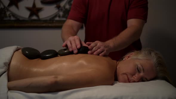 Medium tight shot of a mature caucasian topless woman getting a massage by a man running the stones