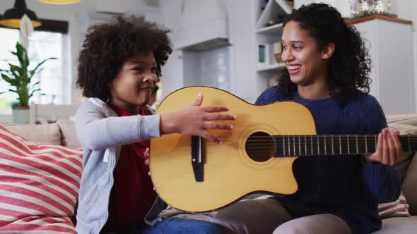 Mixed race mother and daughter sitting on couch playing guitar