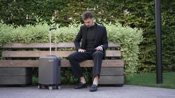 Positive Young Businessman Sitting in City Park with Baggage Checking Time and Departure Ticket with