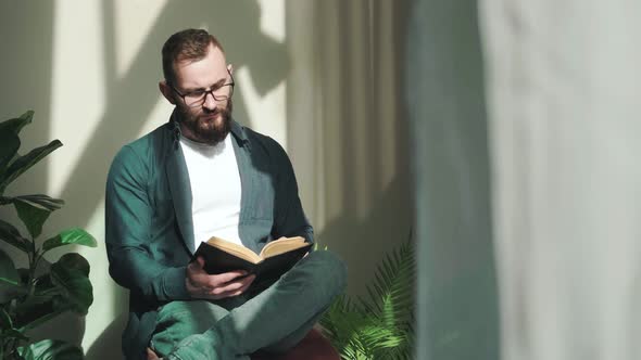 Bearded Young Man Sitting Near Window and Reading a Book at the Sunrise