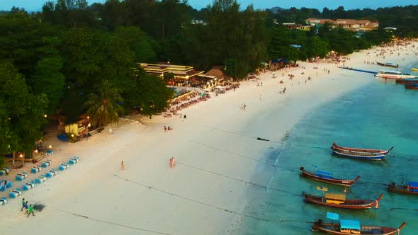 An aerial footage of long white sand beach with benches, unrecognisable people and long tail boats p
