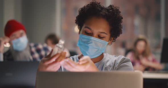 Mixed Race Woman Wearing Face Mask Sanitizing Hands at Desk in Office