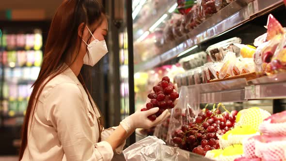 Beautiful attractive asian woman wearing medical face mask and rubber gloves choosing grape on fruit