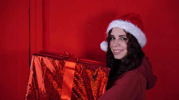 Young Woman in Santa Claus Hat Holds Big Gift Standing Near Red Wall