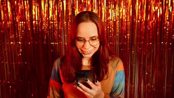 Young Woman in Glasses Browsing Mobile Phone on Background of Shiny Tinsel