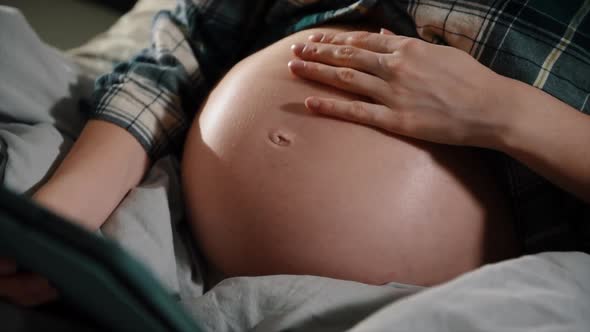 Pregnant with tablet computer in bedroom showing and touching belly.