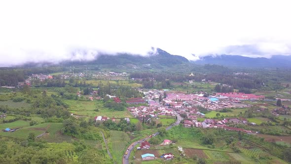 Forward flying aerial view of a beautiful village with green field and mountains covered by the clou