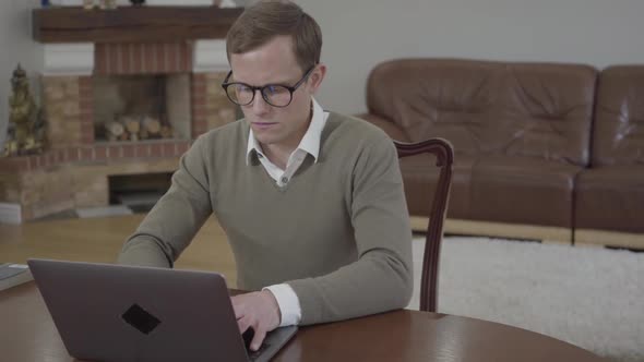 Young Modestly Dressed Man in Glasses Sitting at the Wooden Table in the Office Working