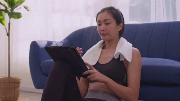 Asian woman in sportswear wiping forehead with towel and using digital tablet while resting after wo