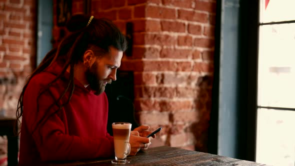 Hipster is Resting in Coffee Shop Drinking Latte and Surfing Internet By Mobile Phone