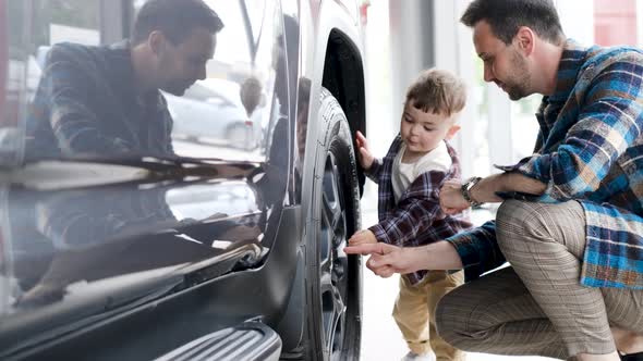 The Happy Owner of the New Car Inspects It in the Showroom with His Young Son