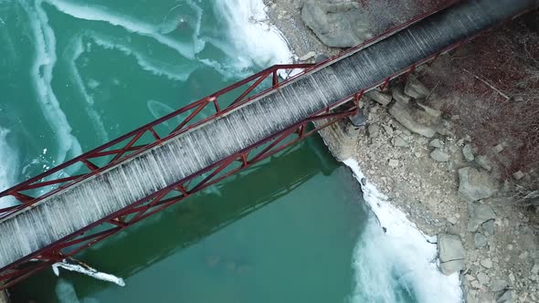 Rusty Footbridge over Icy Mountain River - Top/Drop Drone Shot