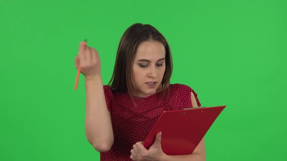 Portrait of Tender Girl in Red Dress Fills Papers in Red Folder with Pensil. Green Screen