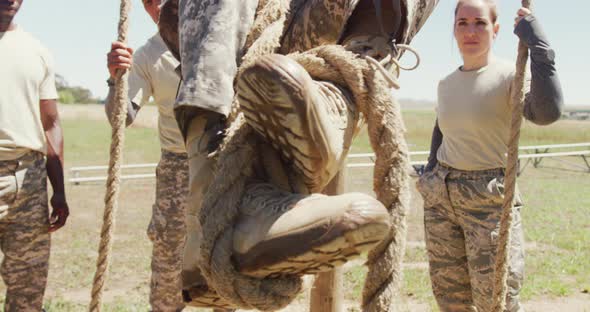 Diverse group watching male soldier in uniform climbing rope on army obstacle course in sun