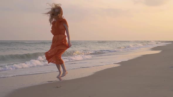 The Young Red-haired Smiling Girl Run in the Orange Dress at Beach By the Sea in Summer.