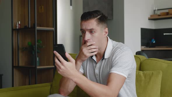 Shocked Man Reacting Message on Smartphone Covers His Mouth in Fright Sitting on Sofa at Home Living