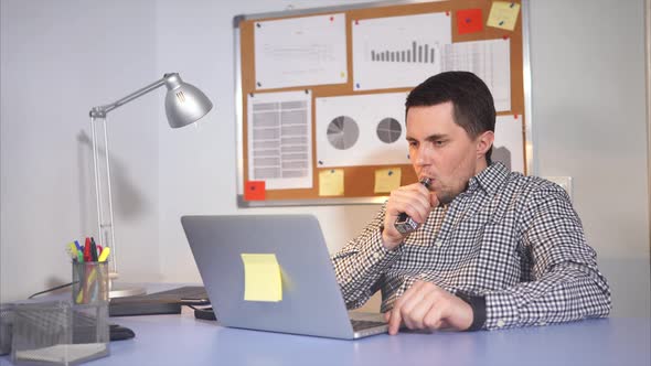 Young Businessman at Work Using Laptop. He Vaping Electronic Cigarette.