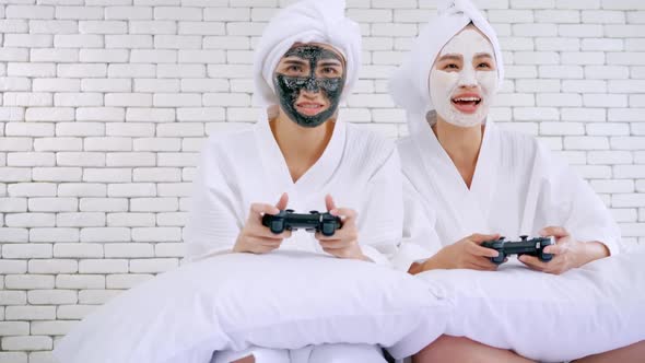 Two happy Asian girls in white bathrobes with facial mask playing video game together in bedroom