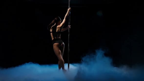 Dancer in a Dark Studio Twirl on a Pole in Blue Smoke. Black Smoke Background. Slow Motion