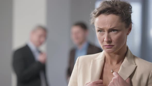 Stressed Brunette Woman with Hazel Eyes Standing Indoors Thinking with Blurred Men Talking at