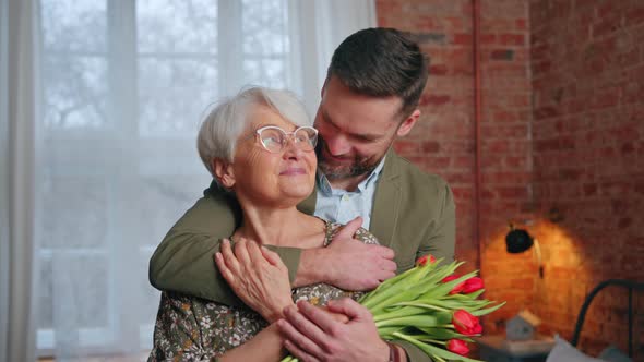 Medium Shot of Elderly Happy Grandma with Her Middleaged Grandson Holding Flowers and Celebrating