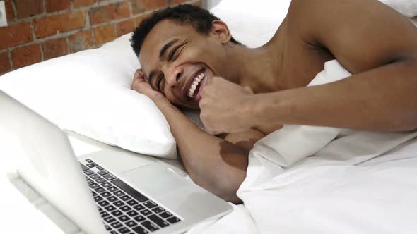 African Man in Bed Working on Laptop and Reacting to Success