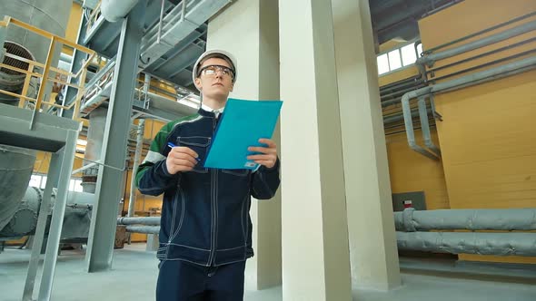 Engineer in Hardhat Walking in Factory and Writing Down on Clipboard