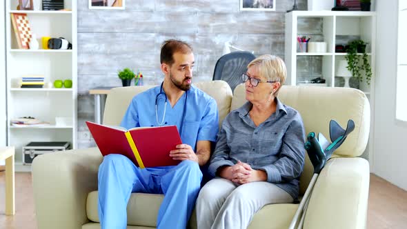 Male Assistant in Nursing Home Reading a Book To Retired Elderly Pensioner