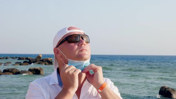 Man in Protective Mask and Sunglasses, Takes Off Mask and Breathes Fresh Air, Against Sea Background