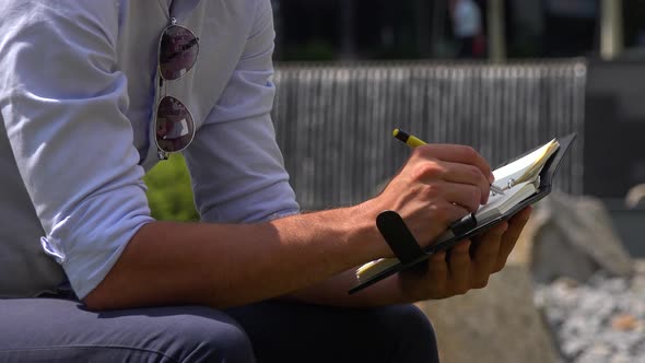 A Businessman Sits in a Park and Writes Notes, Closeup, a Street and Artificial Waterfalls