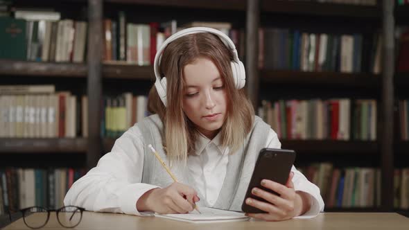 The Schoolgirl with Headphones Teaches Lessons While at Home Sitting in the Kitchen at the Table