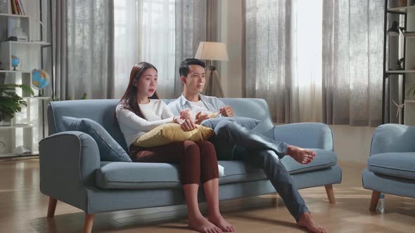 Young Asian Couple Eating Potato Chips And Watching Tv On Couch In Living Room