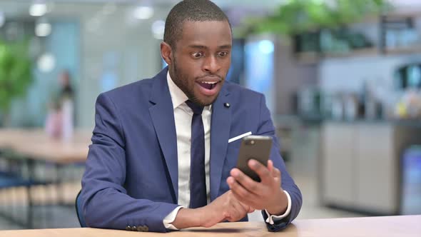 African Businessman Celebrating Success on Smartphone in Office