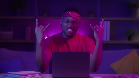 Black Millennial Guy At Laptop Showing Rock Gesture Sitting Indoors