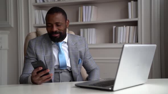 Happy African-American Bearded Man in a Gray Suit, White Shirt. The Businessman Is on the Desktop at
