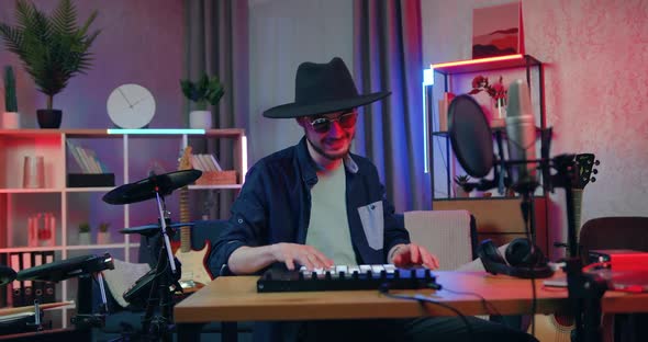 Bearded Guy in Hat and Glasses which Sitting at His Workplace in Home Studio and Playing