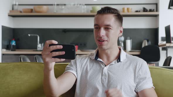 Smiling Young Man with Smartphone Making Video Call While Sitting on Couch in Living Room. Portrait