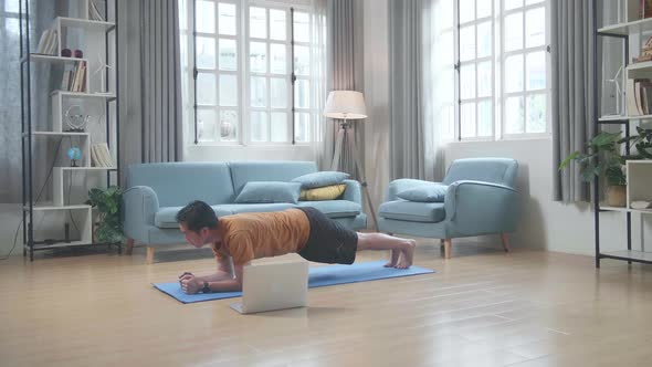 Asian Man On Mat Looking At A Laptop And Doing Yoga In Forearm Plank Pose At Home