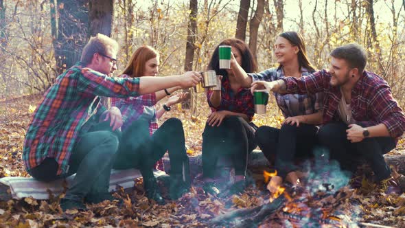 Young hikers toasting with thermo mugs by campfire in autumn forest