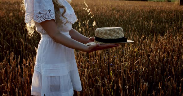 The Girl in a White Dress Puts on a Hat on Her Head on Sunlight in Wheat Field at Incredible