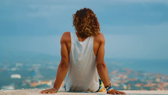 SLOWMO, a young man sits overlooking a coastal city as the wind blows his hair and tank top.Willems