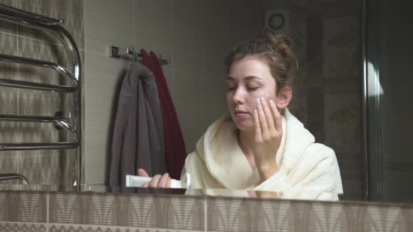 Attractive Young Caucasian Woman in the Bathroom in Front of a Mirror Applies Cream to Her Face