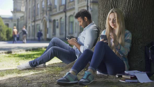 Young Man, Woman Sitting Under Tree With Cellphones, Casting Looks at Each Other