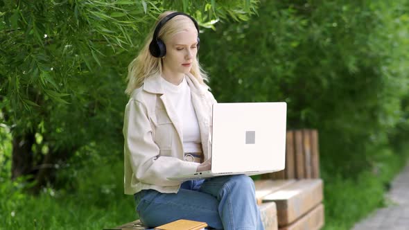 Young Beautiful Blonde Woman in Headphones Sits on a Park Bench with a Laptop